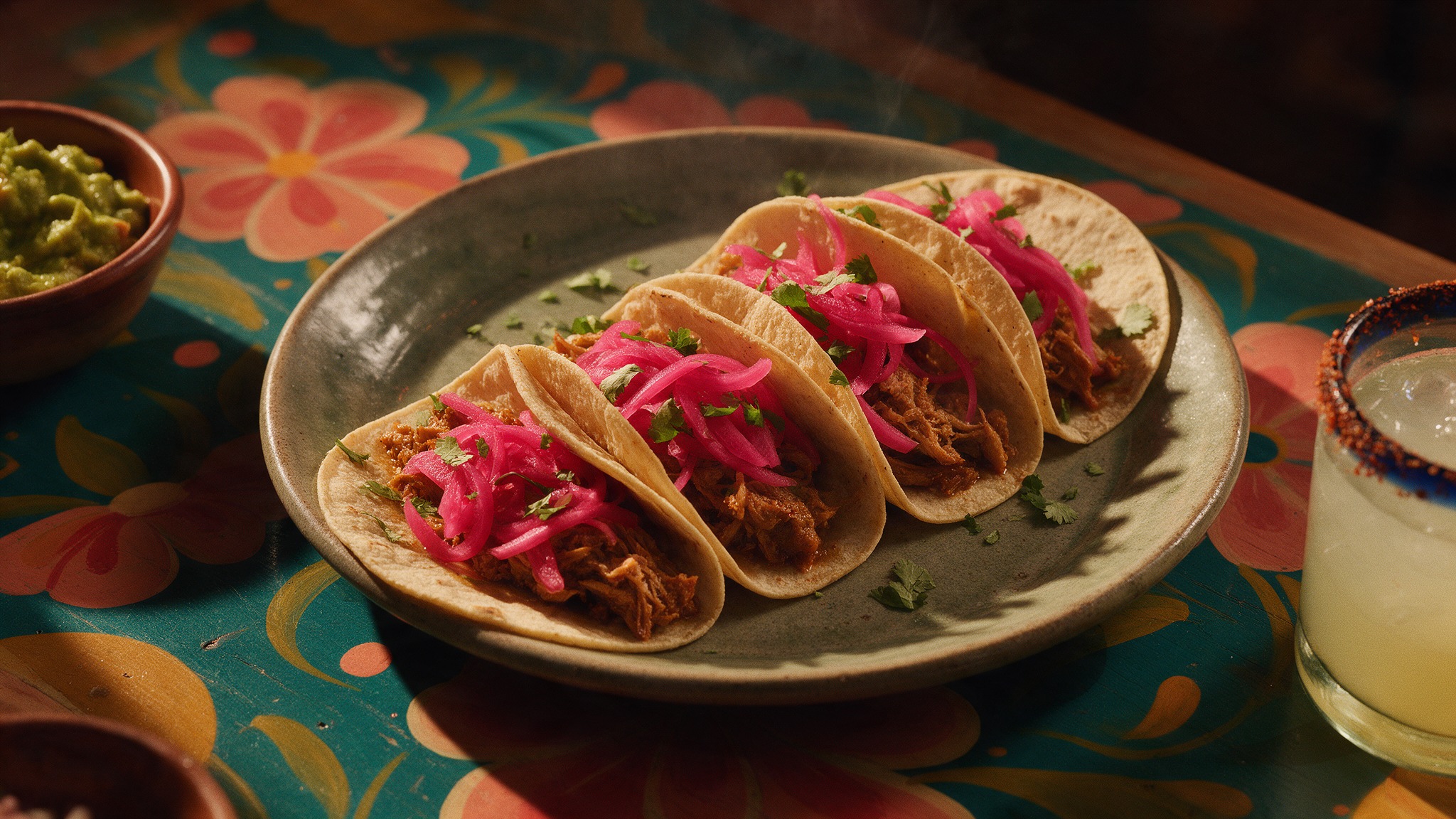 Tacos de cochinita con cebolla encurtida y cilantro, con guacamole y una margarita, sobre una mesa pintada de flores