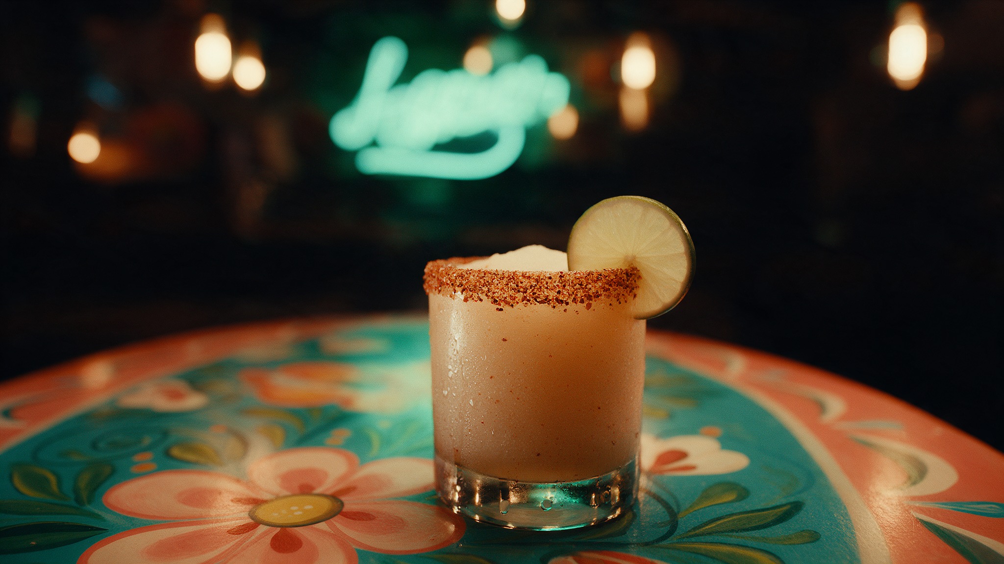 Margarita con borde de chile y sal sobre una mesa pintada, con el neón de la sala al fondo